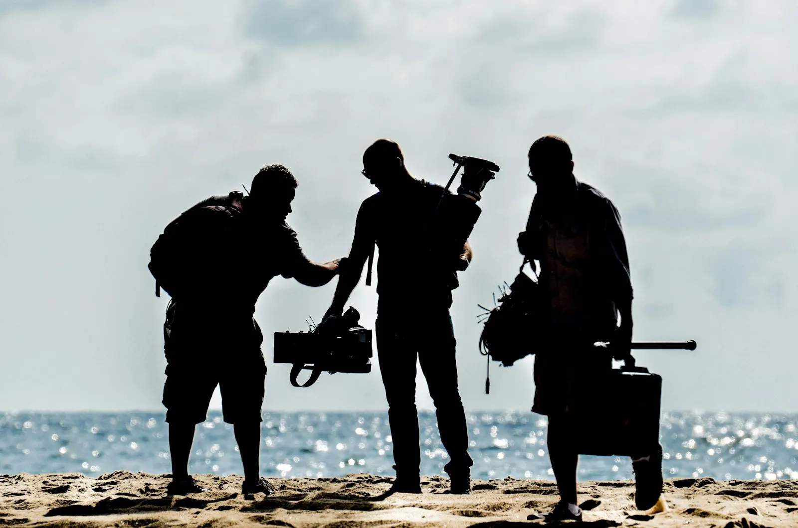 Three crew members silhouetted on a beach carrying camera and sound gear