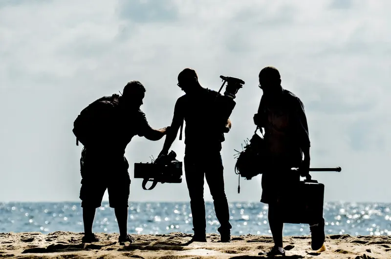 Three camera crew silhouetted on a beach with their kit
