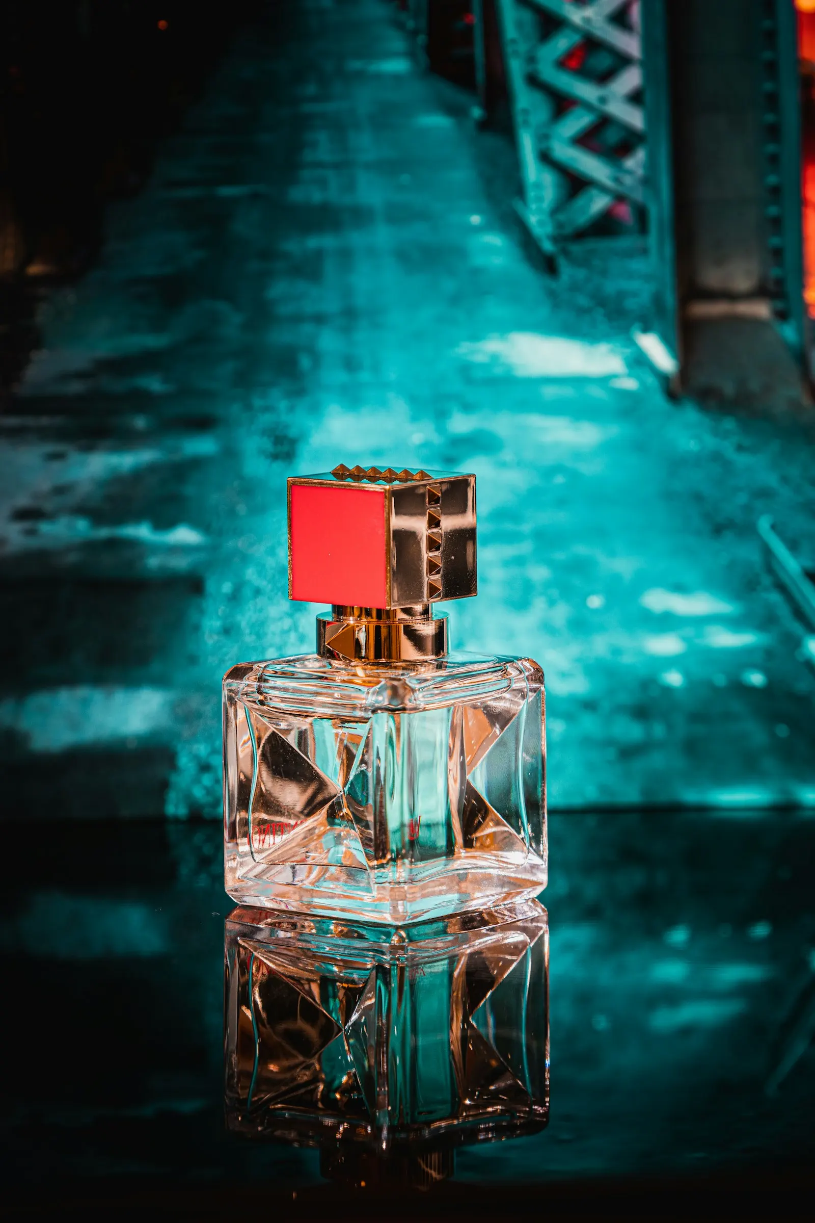 Perfume bottle with a red cap reflected on a wet, teal lit surface